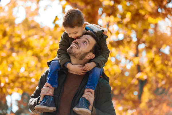 Man carrying a young boy on his shoulders outdoors in autumn with orange leaves in the background.