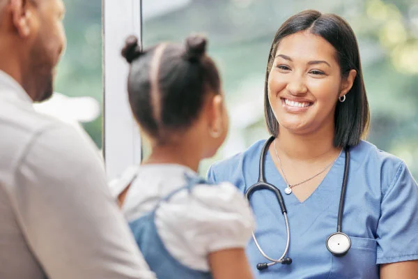 Nurse in blue scrubs smiling while talking with a patient and a child by a window, stethoscope visible.