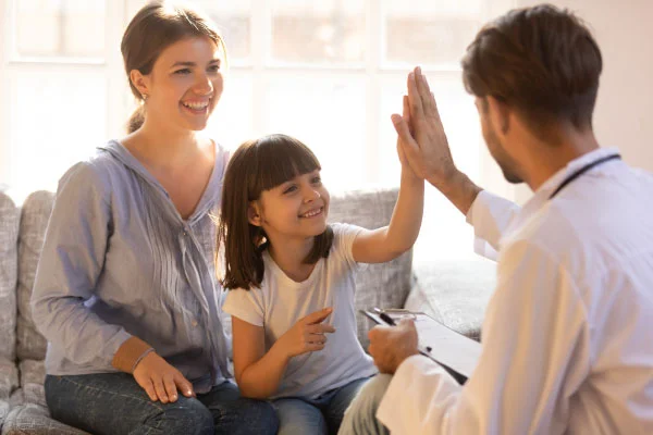 Doctor in a white coat high-fives a smiling young girl seated on a couch, with her mother beside them in a bright room.